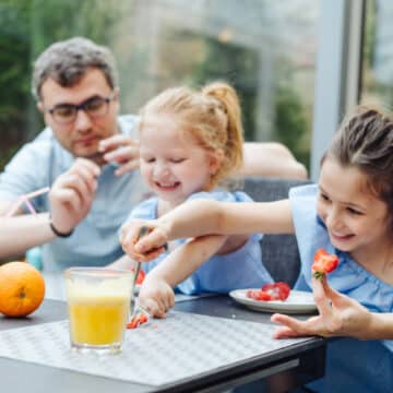 A happy family enjoys a fresh fruit breakfast, with two girls preparing strawberries and an adult overseeing, surrounded by orange juice and whole oranges.