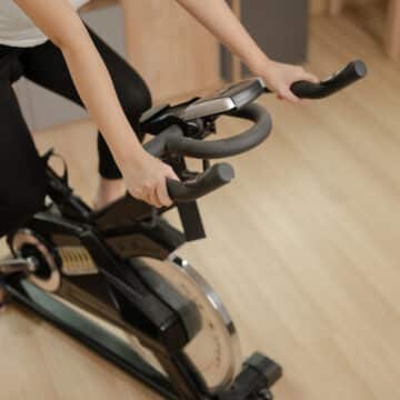 The image shows a woman in sportswear using a stationary bike for indoor cardio, with her hands gripping the handlebars. The room has a wooden floor