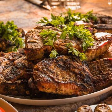 The image shows a plate of grilled meat garnished with parsley. The meat is dark and slightly charred, with some cuts showing bones. The warm, rustic setting suggests a hearty, communal meal