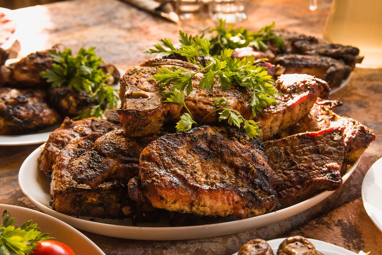 The image shows a plate of grilled meat garnished with parsley. The meat is dark and slightly charred, with some cuts showing bones. The warm, rustic setting suggests a hearty, communal meal